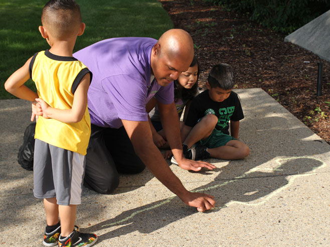 Outside, a student stands, while a teacher traces his shadow with chalk on the sidewalk.