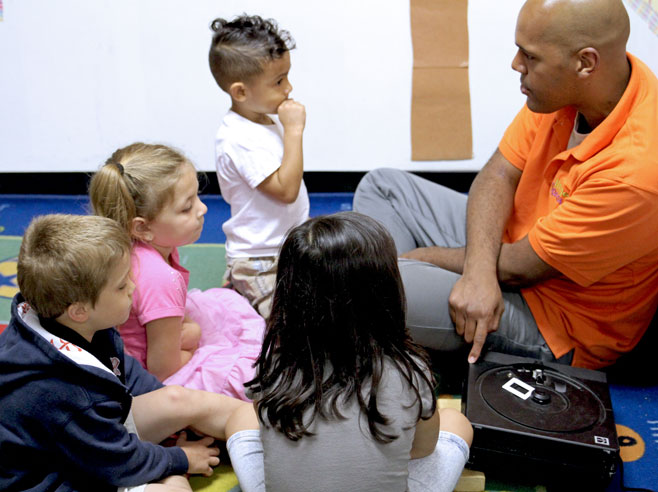A teacher and students are seated on the floor. The teacher is talking and pointing at a projector.