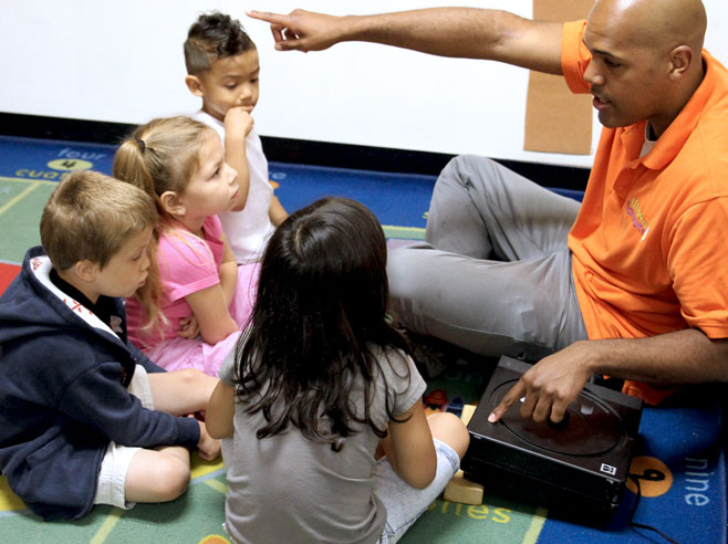 A teacher and students are seated on the floor. The teacher is talking and pointing at a projector.