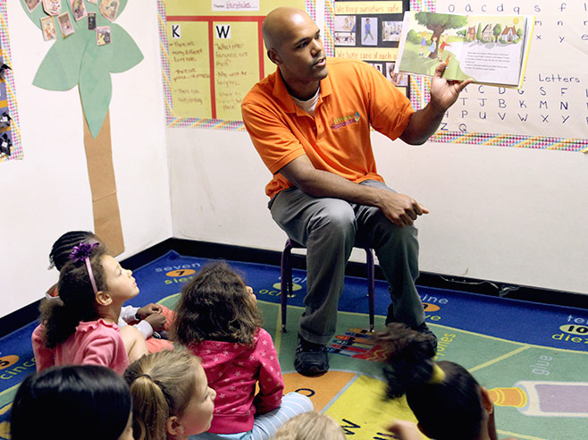 A teacher sits on a chair and holds up a book, open to show illustrated pages. Students are gathered around on the floor as he reads.