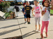 Four students are outside, lined up on a sunny sidewalk. They are jumping, and their shadows are visible.