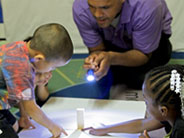 A piece of white poster-board lies on the floor with a wooden block on top. A teacher shines a flashlight at the block, and one student places a craft stick by the block.